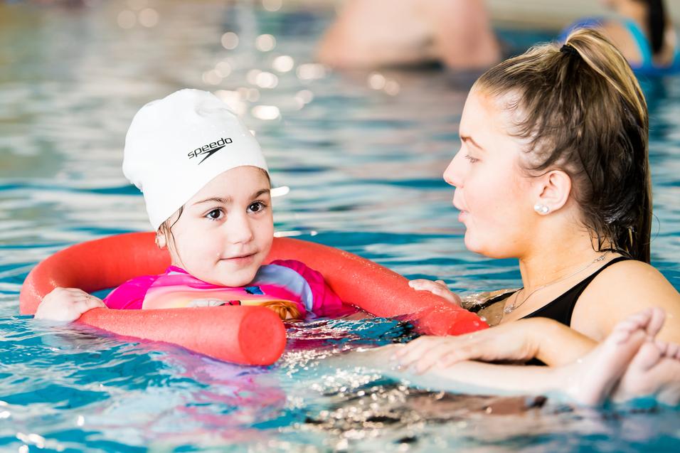Kind mit Schwimmhilfe im Wasser, unterhält sich mit einer Erwachsenen, beide lächeln. Hintergründe zeigen andere Schwimmer.